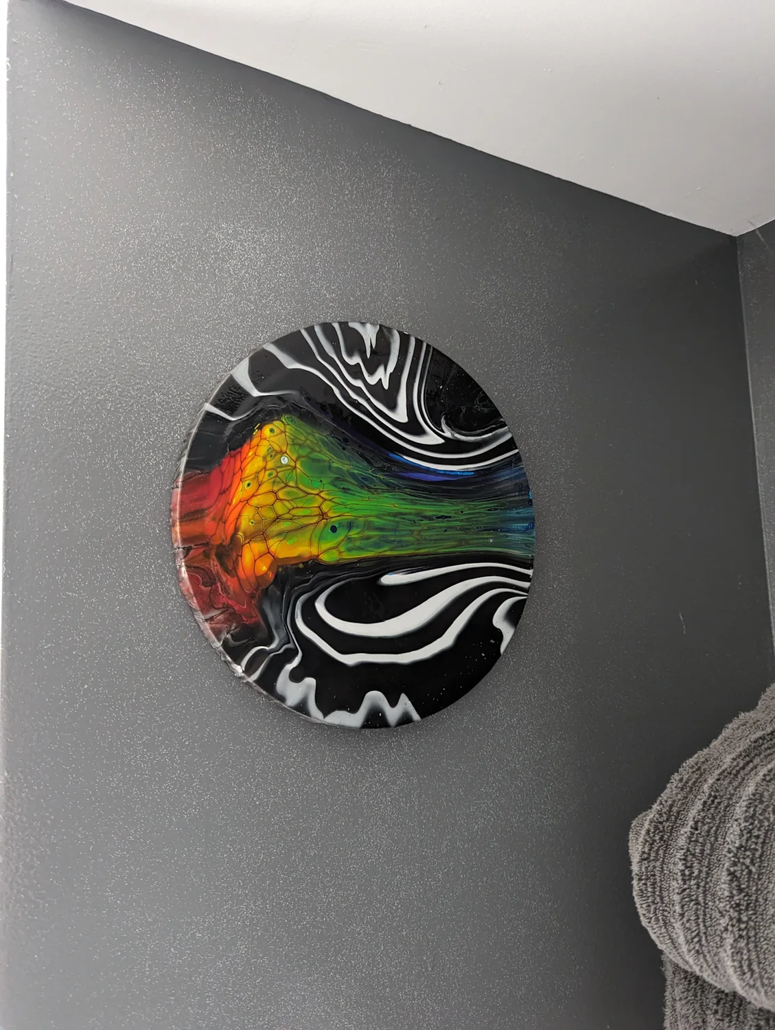A circular black, white and rainbow painting displayed on a gray wall.